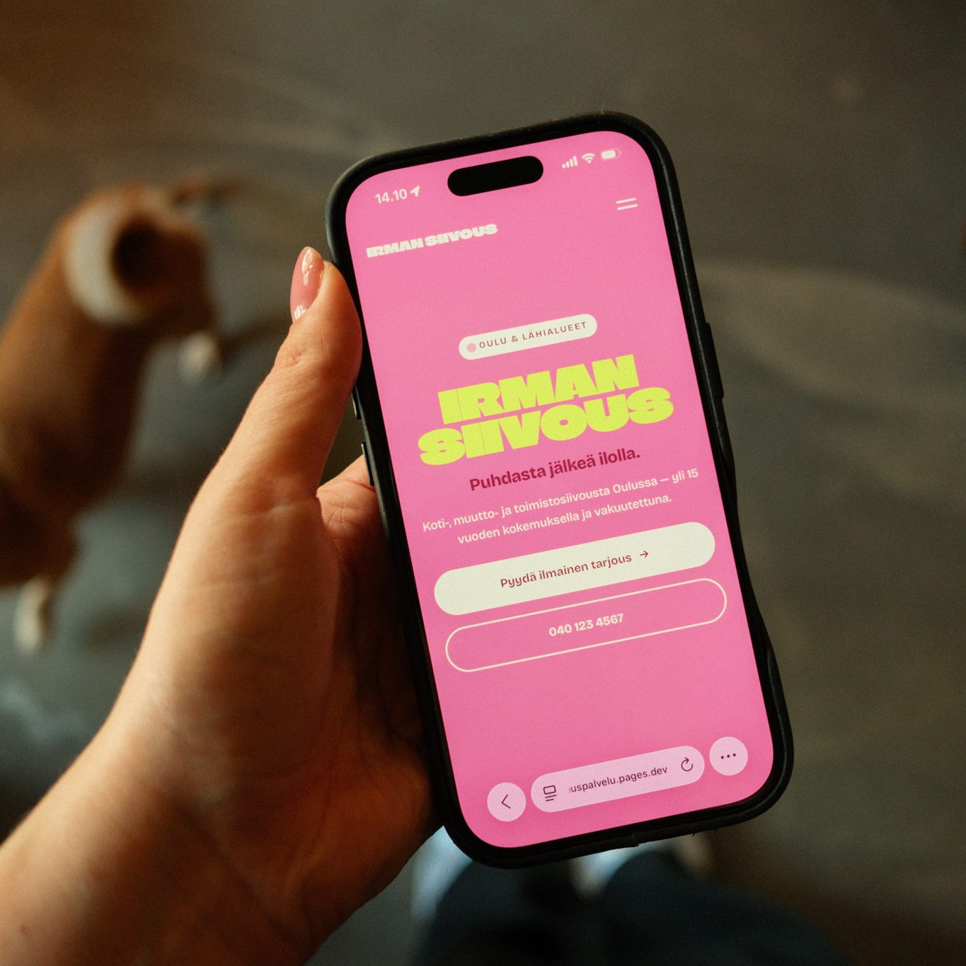 Käsi pitelee puhelinta, jonka näytöllä on Irman Siivouspalvelun pinkki etusivu; taustalla koira.
